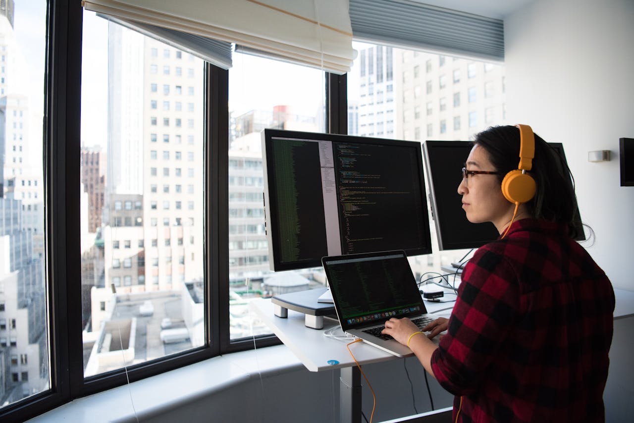 services-03 A businesswoman coding at a standing desk with city views through large windows, wearing headphones.